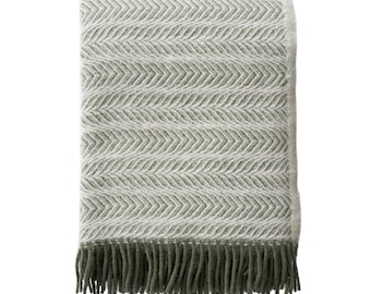 May include: A folded, rectangular blanket with a white and olive green chevron pattern. The blanket has a fringe of olive green tassels along the bottom edge. The blanket is made of a soft, woven material.