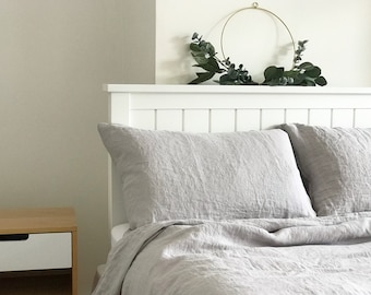 May include: A white bed frame with a gray linen duvet cover and two matching pillowcases. The headboard has a white wood slat design. A gold metal hoop with green eucalyptus leaves is hanging on the wall above the bed.