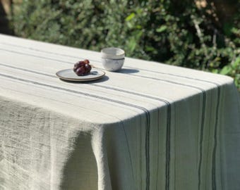 May include: A white linen tablecloth with thin black stripes. The tablecloth is laid out on a table with a small plate of grapes and two white ceramic cups.