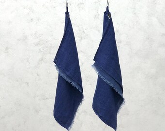 May include: Two indigo blue linen towels with fringed edges hang from metal hooks against a textured, light gray background. The towels are folded and appear to be made of a soft, lightweight fabric.