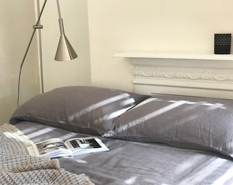 May include: A gray linen bedding set with two pillows and a blanket. The bedding is on a bed with a white headboard and a lamp on the left side of the bed.