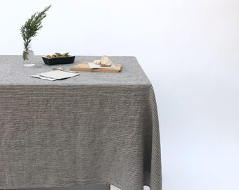May include: A black and white striped linen tablecloth with a small wooden cutting board and a black bowl with olives on top. The tablecloth is draped over a white table.