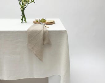 May include: A white linen tablecloth with a beige linen napkin folded on top. A wooden serving tray with crackers and grapes sits on the table. A vase with white flowers sits on the table.