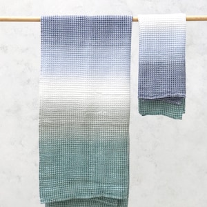 Op de afbeelding: Twee wafelgeweven handdoeken in blauw, wit en groen hangen aan een houten stok. De grotere handdoek heeft een kleureffect, van blauw naar wit naar groen. De kleinere handdoek weerspiegelt het kleurenschema.