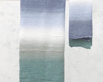 May include: Two waffle-weave towels in shades of blue, white, and green hang from a wooden rod. The larger towel has a gradient effect, transitioning from blue to white to green. The smaller towel mirrors the color scheme.