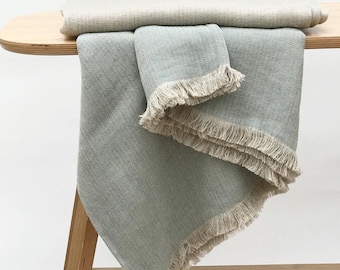 May include: A soft, light green throw blanket with a herringbone pattern and a fringe border. The blanket is folded and draped over a wooden table.