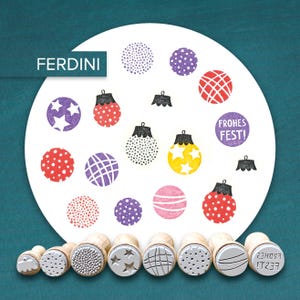 Può includere: Una collezione di timbri in legno con vari disegni, tra cui stelle, punti e motivi a griglia. I timbri sono disposti sotto un cerchio bianco con disegni di ornamenti natalizi stampati in rosso, viola e giallo. Il testo "FROHES FEST!" è anch'esso stampato.