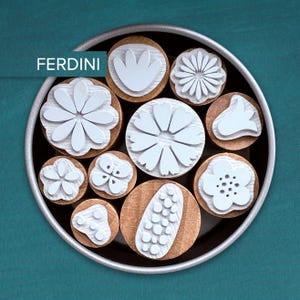 Könnte beinhalten: Eine Sammlung von Stempeln mit floralen Motiven in Weiß auf Holzsockeln, angeordnet in einer kreisförmigen Metallschale. Das Wort "FERDINI" ist sichtbar. Die Stempel zeigen verschiedene Blüten- und Blattformen.