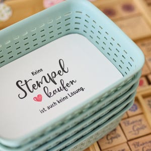 May include: A stack of light blue plastic storage trays. The top tray contains a white card with the words "Keine Stempel kaufen" in black script, a red heart, and additional text. The trays are stacked on top of each other.