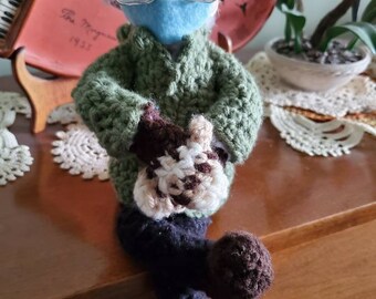 Pode incluir: Uma boneca de crochê vestindo um suéter verde, uma máscara facial azul e óculos. A boneca está segurando um animal de crochê em suas mãos. A boneca está sentada em uma superfície de madeira.
