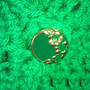 Pode incluir: Um tecido de croch&ecirc; verde com um bot&atilde;o verde e dourado com um design floral.
