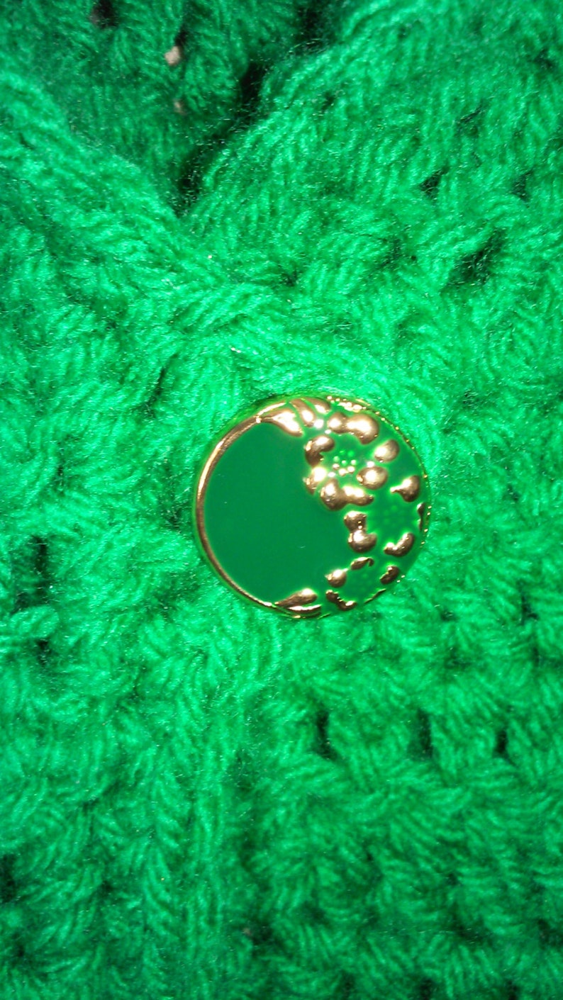 Pode incluir: Um tecido de croch&ecirc; verde com um bot&atilde;o verde e dourado com um design floral.