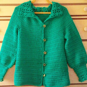 Pode incluir: Um casaco de tric&ocirc; verde com gola e fecho de bot&otilde;es. O casaco tem uma guarni&ccedil;&atilde;o de renda na gola e nas mangas.