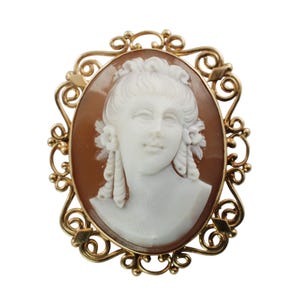 Op de afbeelding: Een gouden cameo broche met een gegraveerd portret van een vrouw met een witte cameo in een gouden filigraan frame.