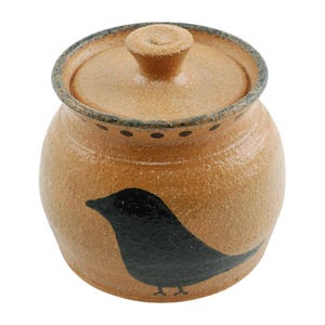 May include: A tan ceramic jar with a lid and a black bird design. The jar has a round shape with a lid and a small knob on top. The bird is painted on the side of the jar. The rim of the jar and lid have a dark blue trim.