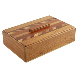 May include: A rectangular wooden box with a lid. The lid features a decorative pattern of inlaid wood in various shades of brown, red, and gold. A small, rectangular handle is centered on the lid. The box is made of light brown wood.