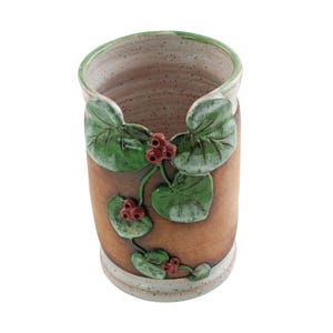 May include: A handmade ceramic vase with a brown exterior and a speckled interior. The vase is adorned with green leaves and red berries, adding a natural touch. The rim and base have a white trim. This decorative piece is ideal for home decor.