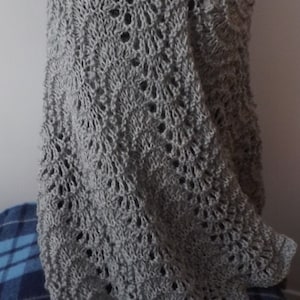 May include: A grey knitted shawl with a lacy pattern. The shawl is draped over a mannequin and has a decorative pin.