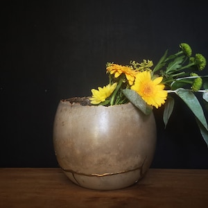 Puede incluir: Un jarrón de cerámica gris agrietado con detalles dorados contiene un ramo de flores amarillas y hojas verdes.