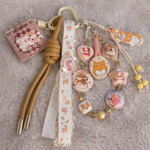 May include: A collection of corgi-themed keychains and accessories. Includes a small notepad with a corgi image, a decorative ribbon, and several charms with corgi illustrations. The color palette is soft, featuring pinks, browns, and creams.