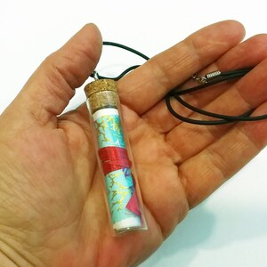 May include: A glass vial necklace with a cork stopper. The vial contains a rolled up piece of paper with a blue and red patterned design. The necklace is made of black cord.