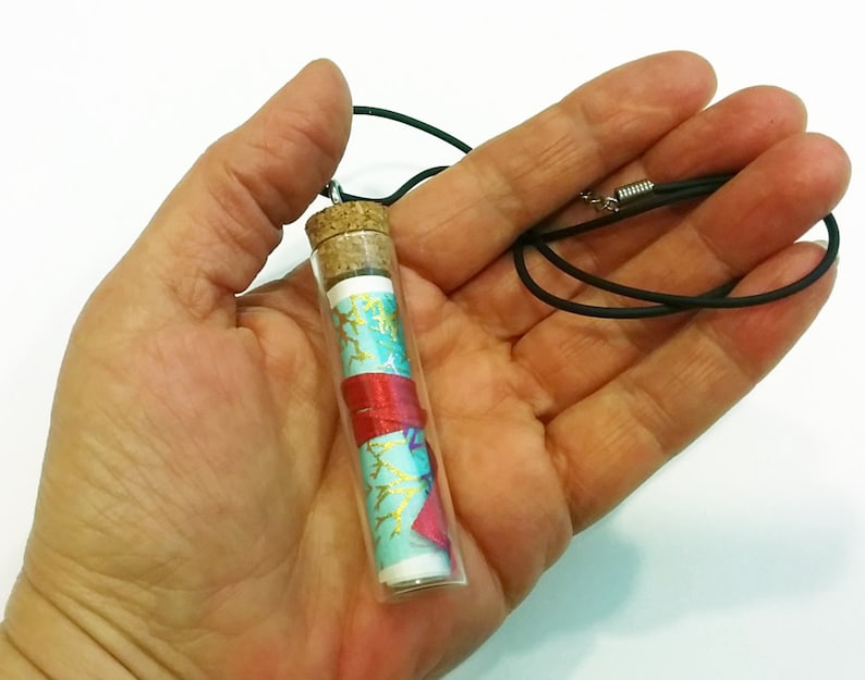 May include: A glass vial necklace with a cork stopper. The vial contains a rolled up piece of paper with a blue and red patterned design. The necklace is made of black cord.