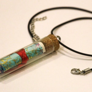 May include: A glass vial necklace with a cork stopper. The vial contains a rolled up piece of fabric with a blue, red, and gold pattern. The necklace is made of black cord and has a silver clasp.