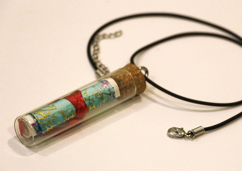 May include: A glass vial necklace with a cork stopper. The vial contains a rolled up piece of fabric with a blue, red, and gold pattern. The necklace is made of black cord and has a silver clasp.