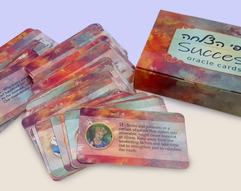 Op de afbeelding: Een set Success orakelkaarten met een rechthoekige doos met een aquarelachtig oranje, rood en geel patroon. De doos toont de tekst Success oracle cards in het Engels en Hebreeuws. De kaarten bevatten cirkelvormige illustraties en tekst.