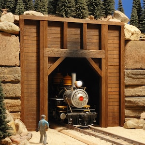 May include: A model train engine emerges from a wooden tunnel entrance. The engine is black with gold accents and has a red number "17" on the side. The tunnel entrance is made of weathered wood planks and has a dark interior.