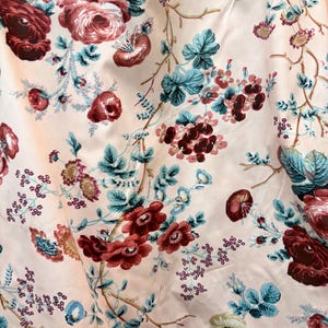 May include: Close-up of a fabric with a floral pattern. The design features large, deep red roses and smaller burgundy flowers, along with teal leaves and delicate blue accents. The background is a soft, pale pink, creating a vintage aesthetic.