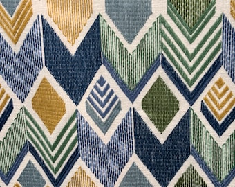 May include: Close-up of a fabric with a geometric pattern. The design features diamond and chevron shapes in shades of blue, green, and gold, set against a cream-colored background. The fabric has a textured appearance, suggesting a woven or embroidered construction.