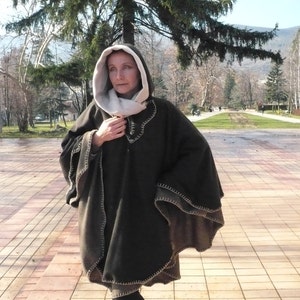 May include: A brown hooded poncho with a cream colored lining and a decorative stitch detail. The poncho is worn by a person standing in a park.