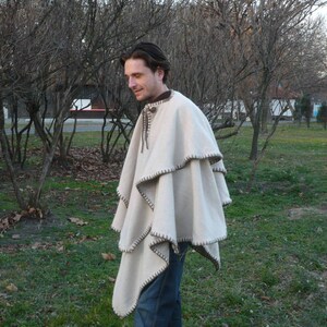 Könnte beinhalten: Ein weißer Wollponcho mit braunem Besatz. Der Poncho hat ein mehrschichtiges Design und wird von einer Person getragen, die in einem Park steht.