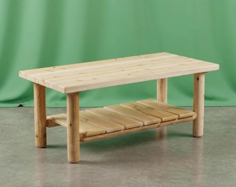 May include: A rectangular coffee table constructed from light-colored natural wood. The table features a slatted top surface and a lower slatted shelf, supported by four sturdy cylindrical legs. The wood grain is visible throughout the piece.