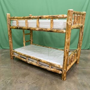 May include: A bunk bed constructed from natural, unfinished logs with visible bark texture. The frame features four vertical corner posts, horizontal side rails, and a built-in ladder on the side. Two mattresses are included, each wrapped in clear plastic.