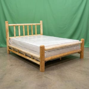 May include: A bed frame constructed from light-colored, unfinished natural logs featuring a headboard with vertical slats and a footboard with a horizontal rail. A white mattress wrapped in clear plastic is positioned on the bed frame.