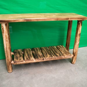 Puede incluir: Una mesa de madera rústica con un solo estante. La mesa está hecha de madera natural con una mancha marrón oscura. La mesa tiene una parte superior rectangular y un estante rectangular.