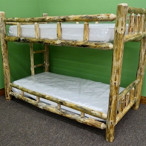 May include: A wooden bunk bed with a rustic, natural finish. The bed is made of thick, rough-hewn logs and has a ladder on one side. The bed is unmade and has white sheets on both mattresses.