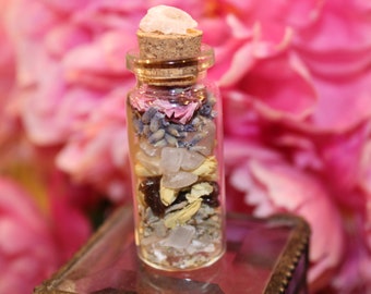 May include: A small glass bottle filled with various crystals, dried flowers, and herbs. The bottle has a cork stopper and is sitting on a glass surface.