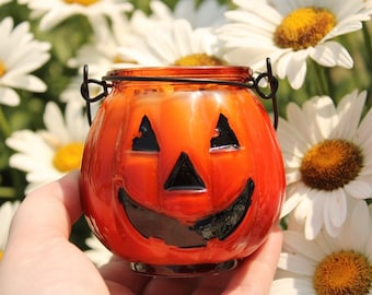 May include: A small orange glass pumpkin candle holder with a black painted face and a wire handle. The candle holder is being held in a hand against a background of white daisies.