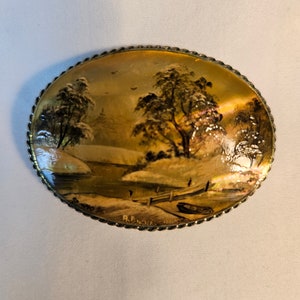 May include: A vintage oval-shaped brooch with a painted scene of a winter landscape. The scene features a river, trees, and a small boat. The brooch is made of metal with a gold finish and has a rope-like border.