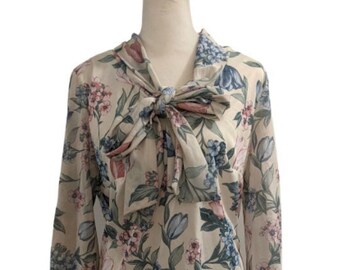 May include: A long-sleeved blouse with a floral pattern in pink, blue, and green on a cream background. The blouse features a tie-neck detail and is displayed on a mannequin.