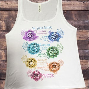 May include: White tank top with a colorful illustration of the seven chakras, including their names and Sanskrit symbols. The chakras are depicted in a watercolor style with floral accents. The text "THE SEVEN CHAKRAS" is at the top of the design.
