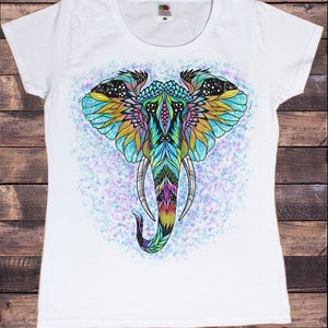 May include: White t-shirt with a colorful, detailed illustration of an elephant's head. The elephant's head is decorated with intricate patterns and colors, including blue, green, yellow, and pink. The background is a light blue with white speckles.