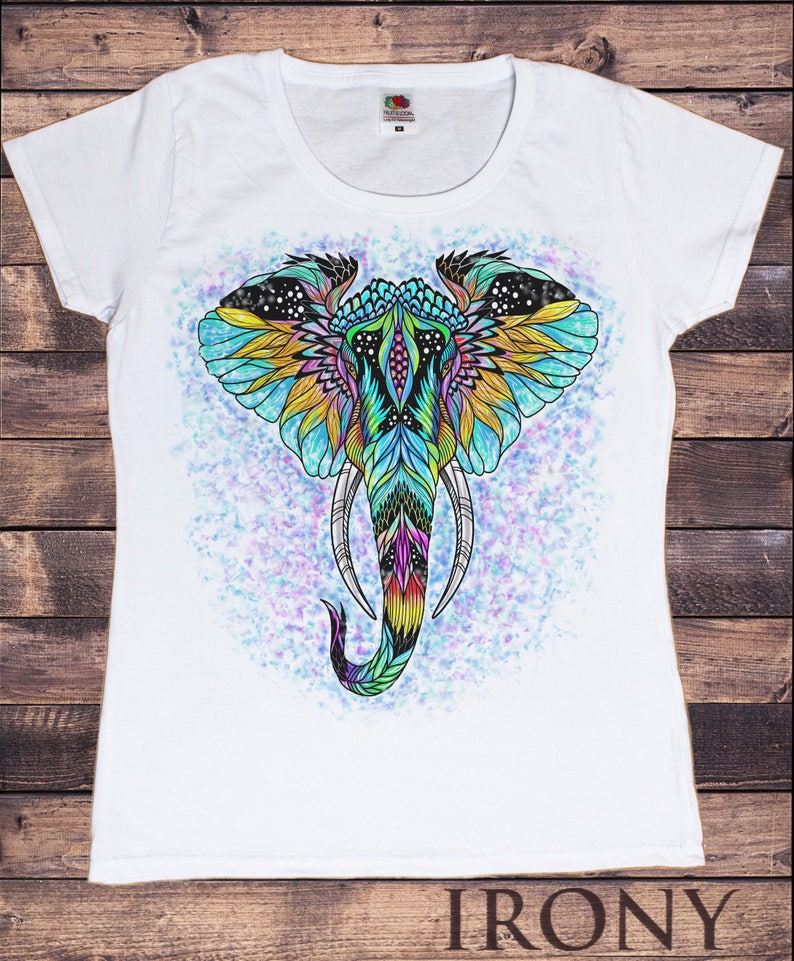 May include: White t-shirt with a colorful, detailed illustration of an elephant's head. The elephant's head is decorated with intricate patterns and colors, including blue, green, yellow, and pink. The background is a light blue with white speckles.