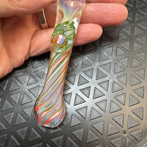 May include: A clear glass pipe featuring a swirling pattern of red, green, and light purple stripes. The pipe includes a decorative glass turtle emblem on the side, which displays a circular design with a red center, yellow border, and blue rays.