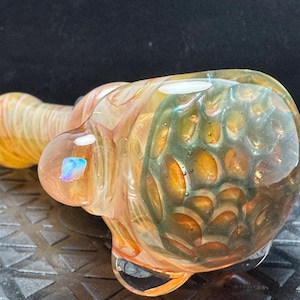 May include: A hand-blown glass pipe with a rounded body and a long, cylindrical mouthpiece. The glass features a blend of orange, yellow, and green hues with a textured, bubble-like pattern. A small, iridescent accent is visible on the surface.