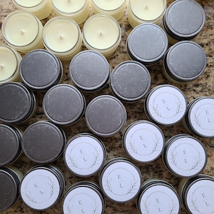 May include: A collection of small glass jars with lids, filled with white candles. The jars have labels with the initials "E" and "L" in a wreath design.