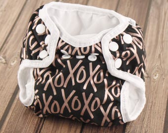 May include: A black and white reusable cloth diaper with a rose gold XOXO print. The diaper has white elastic around the edges and white snaps.
